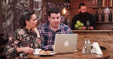 couple en buvant café et à la recherche à ordinateur écran dans très confortable et élégant café boutique, pub ou restaurant avec en bois des murs. une barbu barman est travail dans le Contexte sur le compteur video