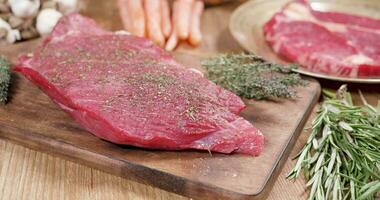glijden over- een vers, rauw steak, goed gekruid Aan een houten bord met tijm en rozemarijn. dik steak en rauw groenten in de achtergrond. video