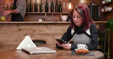 maduro mujer de negocios mensajes de texto en su teléfono y disfrutando un taza de café en Clásico y elegante café tienda video