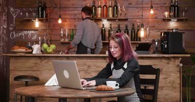 Medium shot of adult businesswoman typing an email while enjoying her coffee. An young bartender is working in the back video