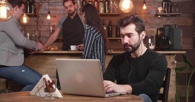 gleiten Schuss von richtig zu links von ein jung gut aussehend Mann Tippen ein Botschaft auf seine Laptop während entspannend im ein Jahrgang Kaffee Geschäft. Barmann und Kunden sind chatten im das Hintergrund. video