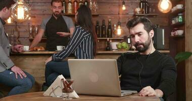 schuif schot van links naar rug van een Kaukasisch mannetje. jong gebaard Mens op zoek Bij zijn laptop scherm terwijl werken van een afstand van een koffie winkel. wijnoogst stijl bezig restaurant. video