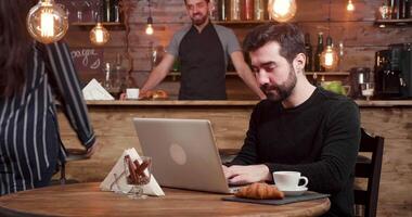 A caucasian man with beard works at his laptop while customers enter the coffee shop. Enthusiastic young businessman works on an online job. video