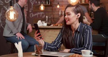 Beautiful female freelancer having a video chat in a coffee shop. Costumers in the background are drinking and chatting wih the bartender.