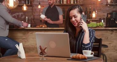 Young pretty lady talking at her phone while working at her laptop remotely from a coffee shop near to her office. Bar in the background. video