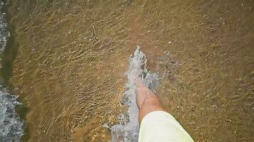 A man walks along the seashore. A view of men's legs walking along the seashore in slow motion and a wave washing over the legs and the sandy shore. video