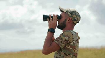 militare osservando attraverso binocolo, uomo nel Camo utilizzando binocolo nel un Aperto campo esamina il del nemico posizione durante dell'ucraina guerra contro Russia video