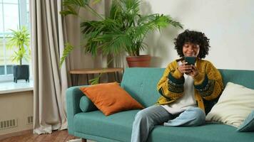 African girl holding smartphone touch screen typing scroll page at home. Woman with cell phone surfing internet using social media apps playing game. Shopping online Internet news cellphone addiction video