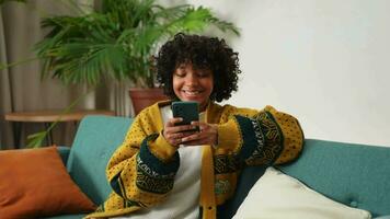 africain fille en portant téléphone intelligent toucher écran dactylographie faire défiler page à maison. femme avec cellule téléphone surfant l'Internet en utilisant social médias applications en jouant jeu. achats en ligne l'Internet nouvelles téléphone portable dépendance video