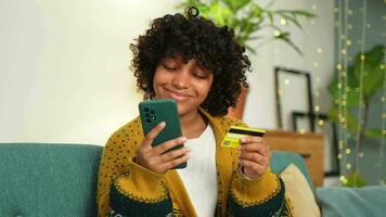 afrikansk amerikan kvinna handla uppkopplad innehav smartphone betalande med guld kreditera kort. flicka Sammanträde på Hem uppköp på internet stiga på kreditera kort detaljer. uppkopplad handla e-handel leverans service video