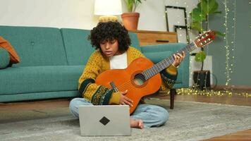 African American woman learning guitar watching tutorial on laptop at home. Black girl playing guitar singing song learning online music lessons. Artistic woman playing acoustic guitar learning chords video