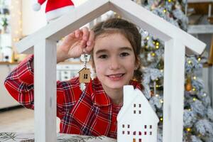 contento niño con minúsculo casa en acogedor hogar con Navidad decoración de festivo blanco cocina. regalo para nuevo año. seguro, Moviente a nuevo casa, hipoteca, alquilar y compra real inmuebles foto