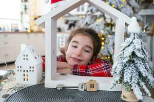 contento niño con minúsculo casa en acogedor hogar con Navidad decoración de festivo blanco cocina. regalo para nuevo año. seguro, Moviente a nuevo casa, hipoteca, alquilar y compra real inmuebles foto