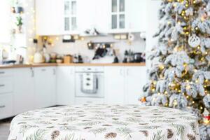 Round empty table to demonstrate the product with with tablecloth with pine needles and cones print, space for text in white kitchen, modern interior with a Christmas tree. New Year, Christmas photo