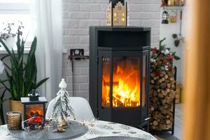 Metall black stove, burning hearth fireplace in white Festive interior of house is decorated for Christmas and New Year, Christmas tree. firewood in the woodpile, cozy and heating of home photo