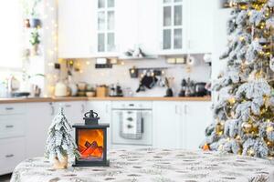 Festive Christmas decor on a round empty table with tablecloth with pine needles and cones print in white kitchen, modern interior with a Christmas tree. New Year, Christmas mood, cozy home. Copyspace photo