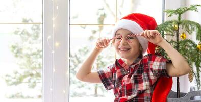 Girl in a Santa hat sits on the windowsill of a house near the Christmas tree and puts Candy cane to her eyes like glasses. Child is having fun and making faces, waiting for Christmas and New year photo