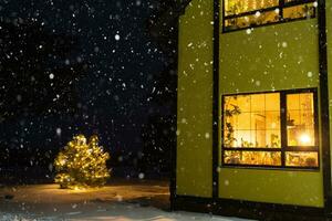 acogedora ventana festiva de la casa exterior con la cálida luz de las guirnaldas de luces de hadas en el interior: celebre la navidad y el año nuevo en un hogar cálido. árbol de navidad, bokeh, nieve en pinos y nevadas foto