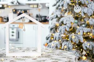 Key and tiny house of a small size on cozy home with Christmas decor on table of festive white kitchen. Gift for New Year. Insurance, moving to new house, mortgage, rent and purchase real estate photo