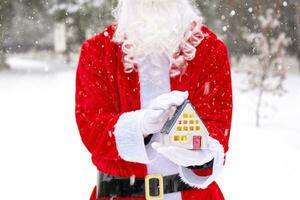 Santa carefully holds in his hands a tiny house in a small size in snow outdoor - a gift for Christmas, New year, the fulfillment of a dream. Protection, insurance, mortgage, purchase of housing photo