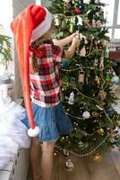 Happy girl in a Santa hat decorates the Christmas tree with colorful figurines. The child is having fun barefoot, in a dwarf's hood near the window, waiting for Christmas and New year photo