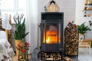 Metall black stove, burning hearth fireplace in white Festive interior of house is decorated for Christmas and New Year, Christmas tree. firewood in the woodpile, cozy and heating of home photo