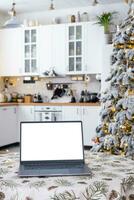 Laptop with a white screen mock up in the cozy white decorated Christmas kitchen with fairy lights and a Christmas tree. Seasonal remote work, internet, shopping, buying gifts Christmas and New Year. photo