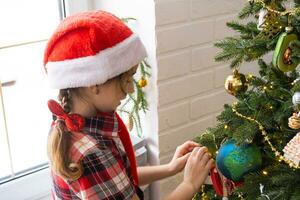 Happy girl in a Santa hat decorates the Christmas tree with colorful figurines. The child is having fun barefoot, in a dwarf's hood near the window, waiting for Christmas and New year photo