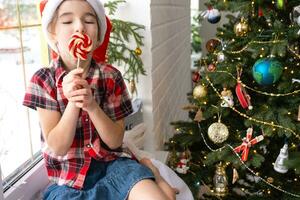 Happy Girl in a Santa hat sits on the windowsill of a house near the Christmas tree and puts sweet round caramel on a stick. Child is having fun and making faces, waiting for Christmas and New year photo