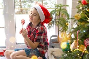 Happy Girl in a Santa hat sits on the windowsill of a house near the Christmas tree and puts sweet round caramel on a stick. Child is having fun and making faces, waiting for Christmas and New year photo