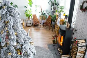 Festive white modern interior of house is decorated for Christmas and New Year in loft style with black stove, fireplace, Christmas tree. Potted plants, firewood in the woodpile photo