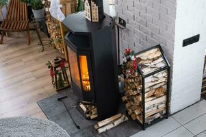 Metall black stove, burning hearth fireplace in white Festive interior of house is decorated for Christmas and New Year, Christmas tree. firewood in the woodpile, cozy and heating of home photo