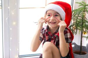 Girl in a Santa hat sits on the windowsill of a house near the Christmas tree and puts Candy cane to her eyes like glasses. Child is having fun and making faces, waiting for Christmas and New year photo