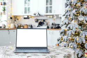 Laptop with a white screen mock up in the cozy white decorated Christmas kitchen with fairy lights and a Christmas tree. Seasonal remote work, internet, shopping, buying gifts Christmas and New Year. photo