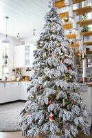 Festive Christmas decor in white kitchen, modern village interior with a snowy Christmas tree and fairy lights. New Year, Christmas mood, cozy home, Metal modular staircase. The general plan photo