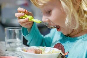 un pequeño niña tiene desayuno a hogar espaguetis con salchichas pequeño rubia niña comiendo cena con tenedor a mesa foto