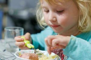 un pequeño niña tiene desayuno a hogar espaguetis con salchichas pequeño rubia niña comiendo cena con tenedor a mesa foto