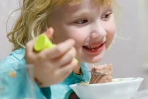 un pequeño niña tiene desayuno a hogar espaguetis con salchichas pequeño rubia niña comiendo cena con tenedor a mesa foto