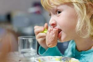 un pequeño niña tiene desayuno a hogar espaguetis con salchichas pequeño rubia niña comiendo cena con tenedor a mesa foto