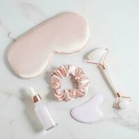 Pink Sleep mask, hair elastic, face roller and essential oil on marble background. The concept of rest, sleep quality, good night, insomnia, relaxation. Personal women accessories photo