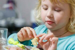 un pequeño niña tiene desayuno a hogar espaguetis con salchichas pequeño rubia niña comiendo cena con tenedor a mesa foto