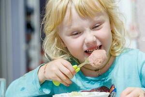 un pequeño niña tiene desayuno a hogar espaguetis con salchichas pequeño rubia niña comiendo cena con tenedor a mesa foto