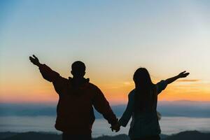 silueta de un romántico Pareja participación manos en parte superior de un montaña a amanecer. foto
