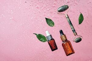 Organic cosmetics, green leaves and jade massage roller. Skincare product bottle and water drops on pink background photo
