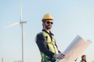 Young man engineer wearing safety helmet and holding blueprint in wind turbine farm. photo