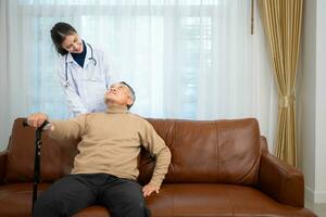 Doctor examines and treats an elderly man seated on a sofa with a cane who is suffering from knee joint pain. photo