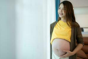 hermosa embarazada mujer en pie cerca ventana a hogar y participación su barriga foto