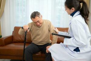 Doctor examines and treats an elderly man seated on a sofa with a cane who is suffering from knee joint pain. photo