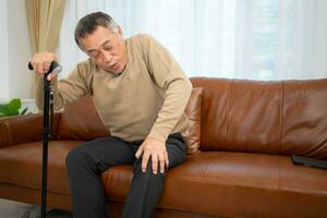 With knee joint pain, a senior guy sits on a sofa with a walking stick in his living room at home. photo