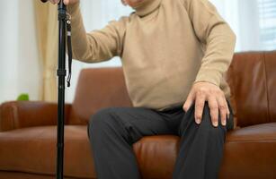 With knee joint pain, a senior guy sits on a sofa with a walking stick in his living room at home. photo